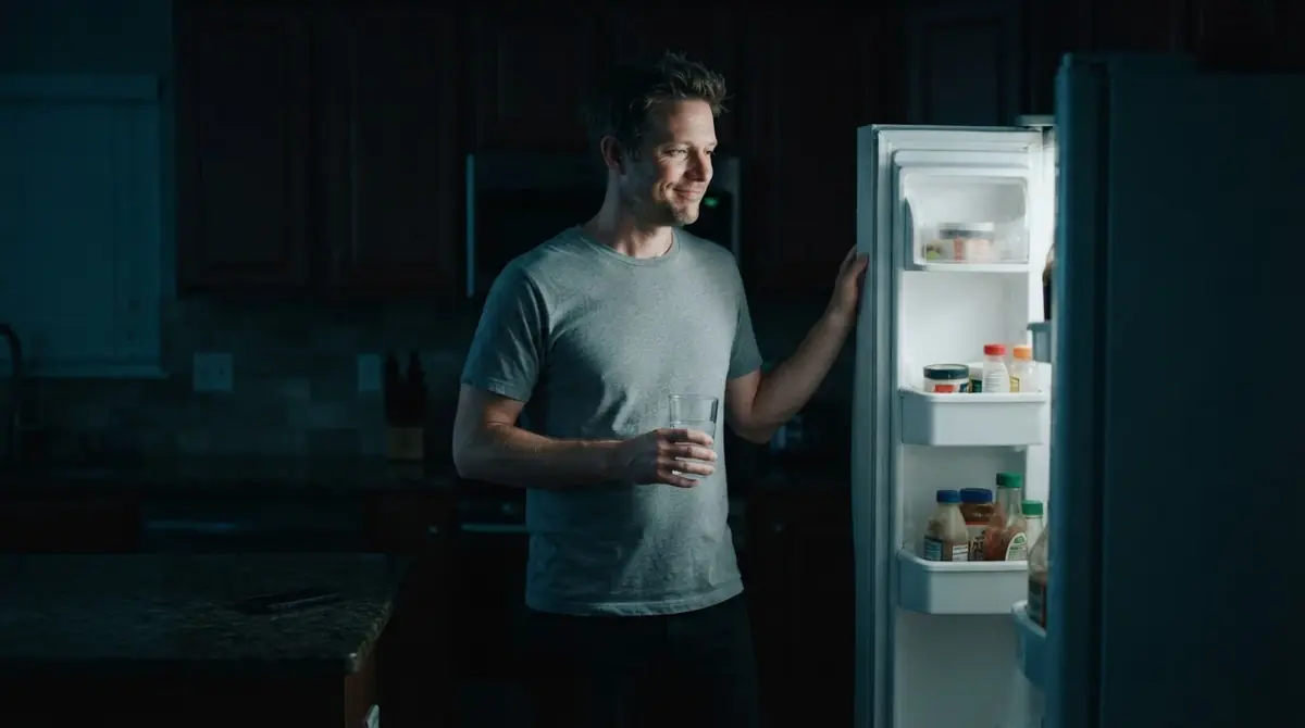 A man closes the fridge in a dark kitchen, holding a glass of water instead.
